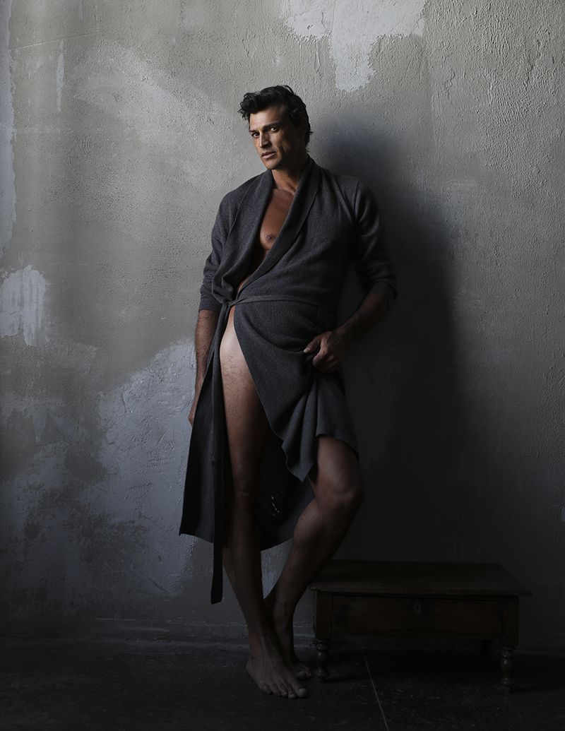 Romulo Pires standing barefoot in an open dark grey bathrobe against a weathered concrete studio wall.