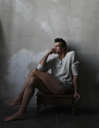Model Romulo Pires seated barefoot on a wooden stool wearing a light ribbed sweater against a textured grey wall
