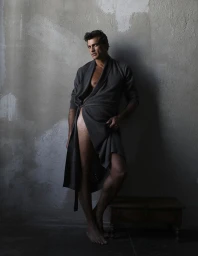 Romulo Pires standing barefoot in an open dark grey bathrobe against a weathered concrete studio wall.