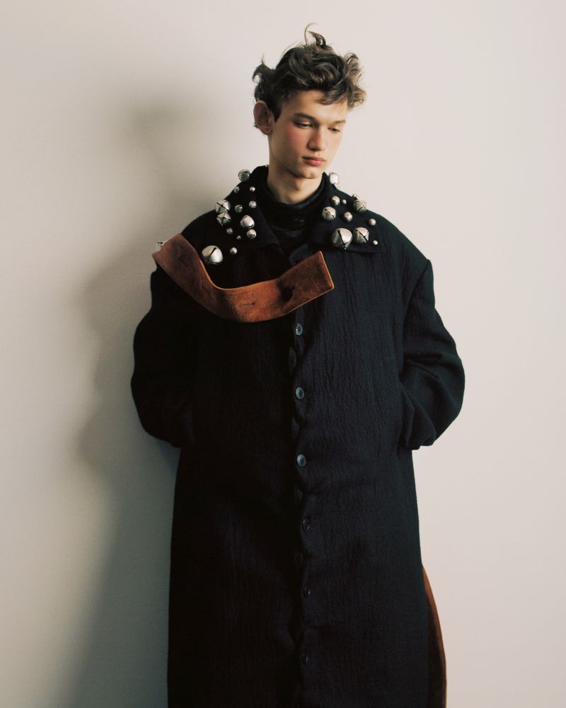 A fair-skinned male model with brown curly hair is wearing a black button-up coat with metal adornments and a brown sash in a studio.