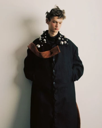 A fair-skinned male model with brown curly hair is wearing a black button-up coat with metal adornments and a brown sash in a studio.