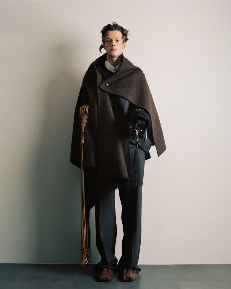 Model wearing a brown asymmetrical draped wool cape and dark tailored trousers for Sean Suen FW25 backstage.