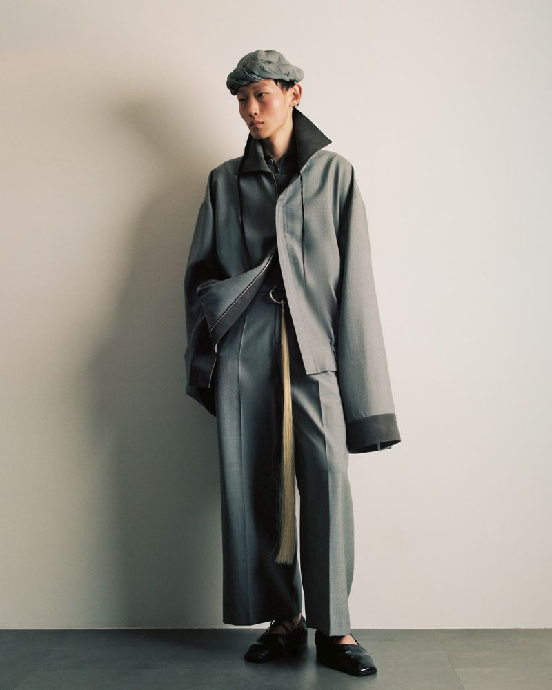 Fashionable individual wearing a gray tailored suit with wide-leg pants and matching cap against a neutral backdrop, in a studio setting.