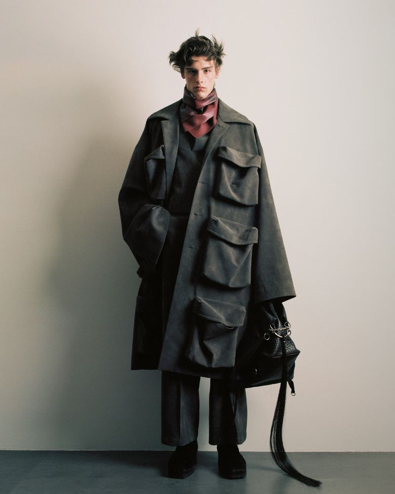 Male fashion model poses in a grey oversized coat with large pockets, accessorized with a paisley scarf and leather handbag against a plain background.