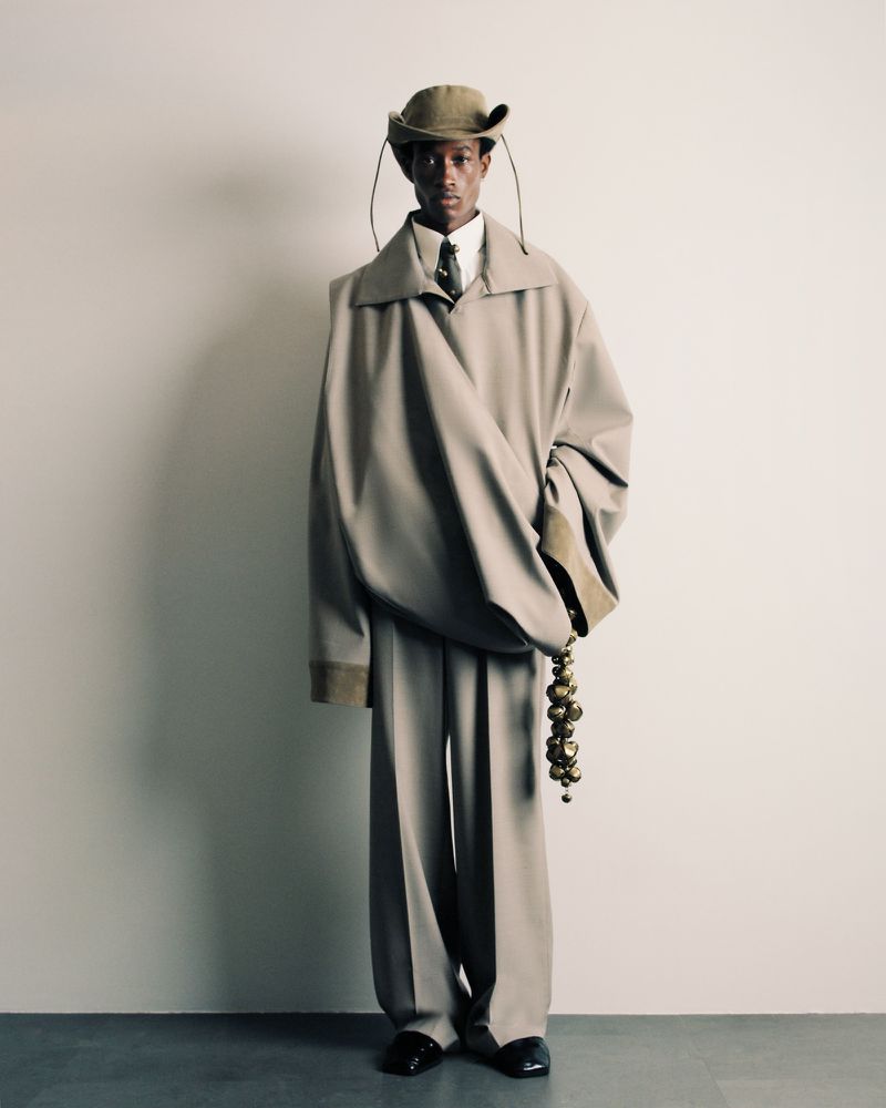 A person in an oversized beige outfit poses in front of a white wall, featuring a jacket, pants, tie, and hat with hanging straps.
