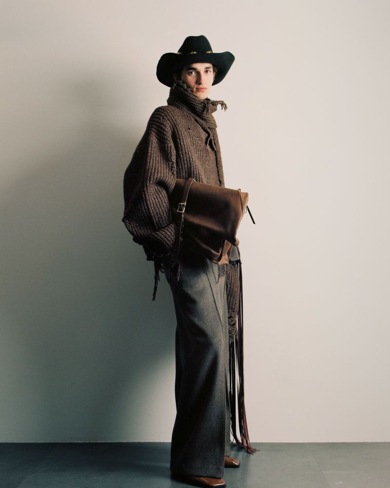 Fashion shot of a model wearing a brown knit sweater, black cowboy hat, and wide-leg pants, holding a leather bag in a studio setting.