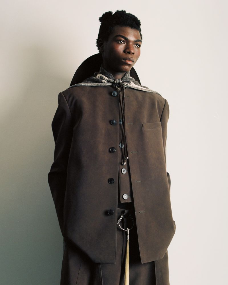 Portrait of a person in a brown suit against a white backdrop, featuring a scarf, neutral colors, and a modern editorial style.