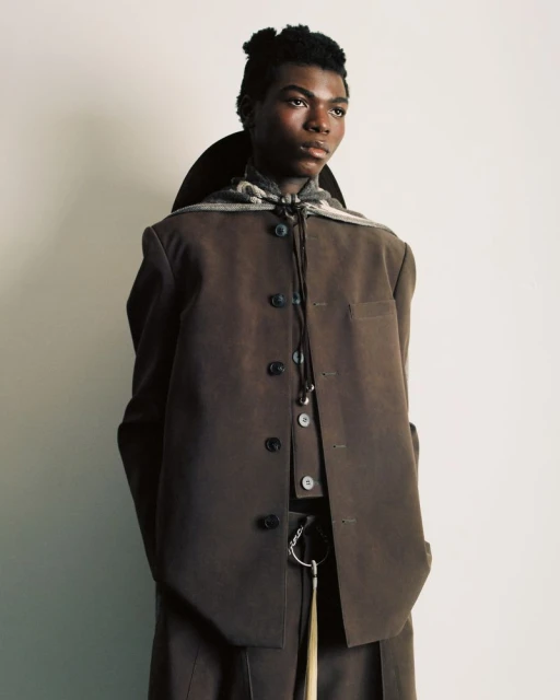 Portrait of a person in a brown suit against a white backdrop, featuring a scarf, neutral colors, and a modern editorial style.