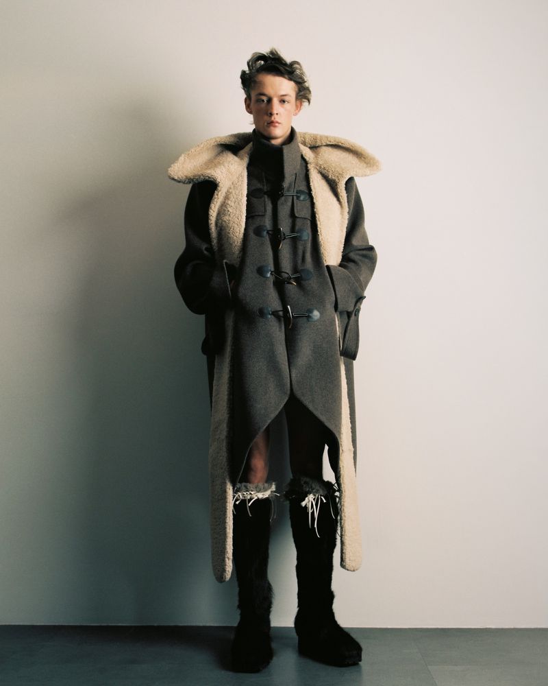 Teen male model poses in an avant-garde fashion outfit featuring a long coat with fur trim and high boots in a studio setting.