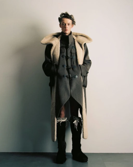 Teen male model poses in an avant-garde fashion outfit featuring a long coat with fur trim and high boots in a studio setting.