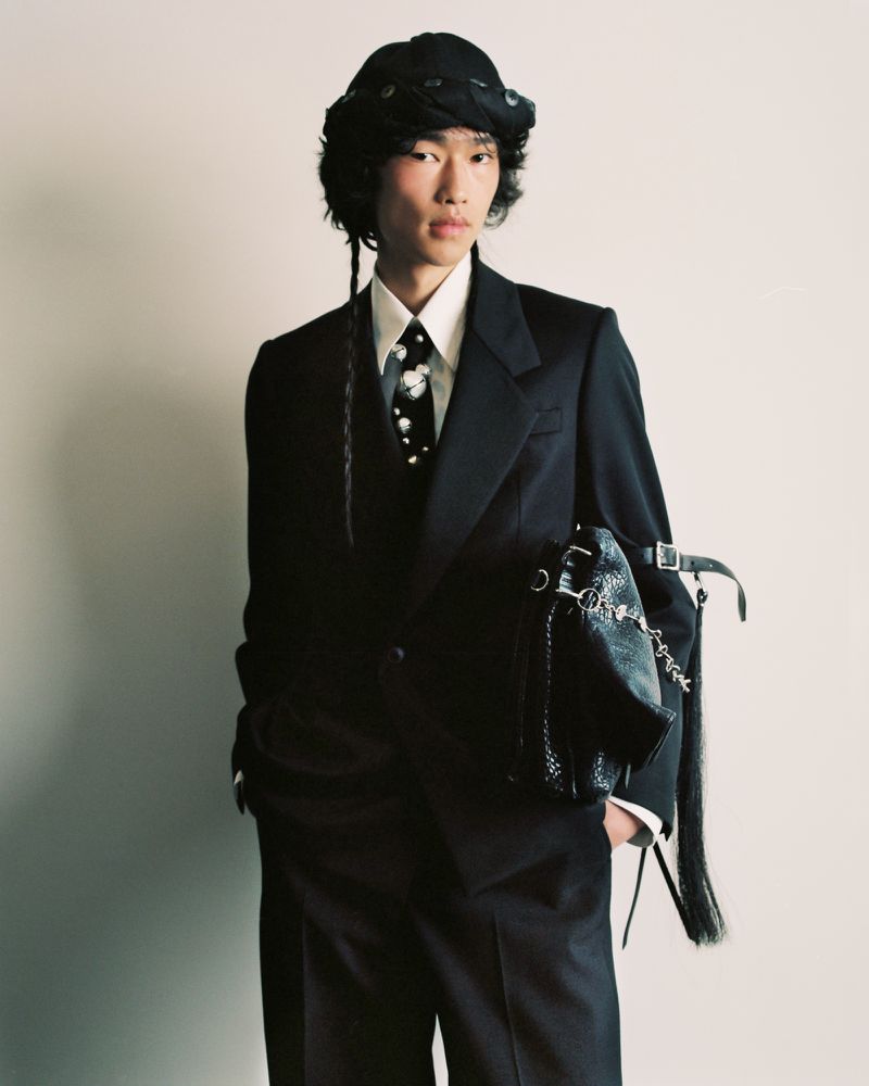 Young Asian male in a dark suit, tie with light floral print, and bucket hat posing with a decorative black bag against a plain wall.
