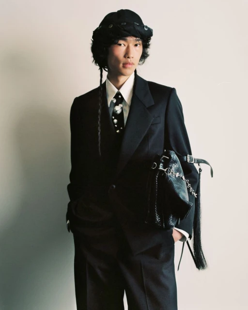 Young Asian male in a dark suit, tie with light floral print, and bucket hat posing with a decorative black bag against a plain wall.
