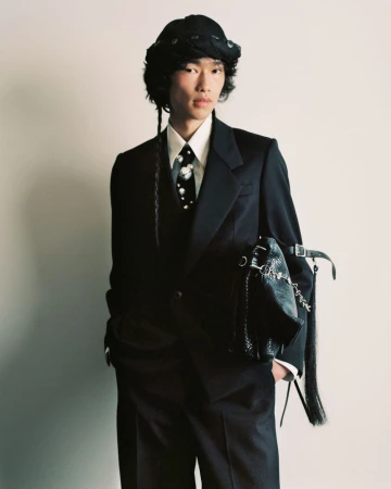 Young Asian male in a dark suit, tie with light floral print, and bucket hat posing with a decorative black bag against a plain wall.