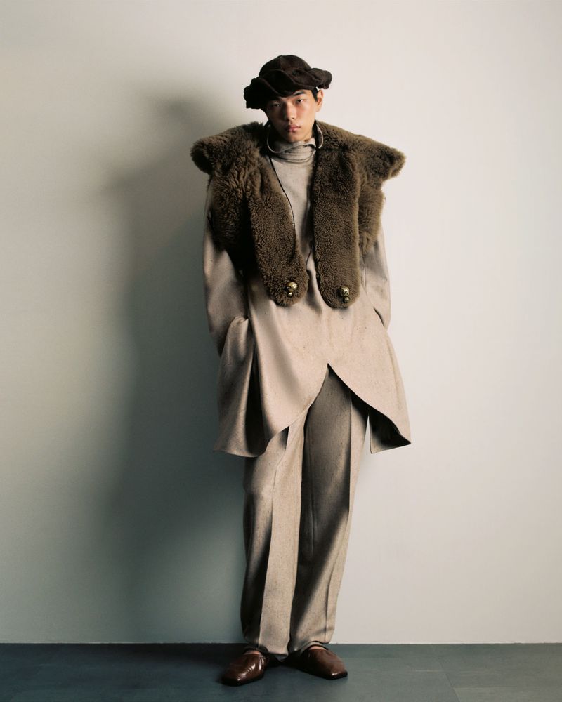Full-length portrait of a person in a stylish beige suit with a fur collar and hat, standing in front of a neutral wall.