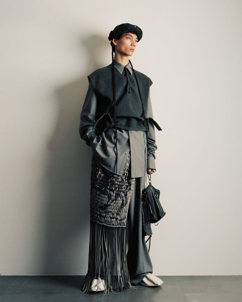 An individual poses in an indoor studio, showcasing a stylish gray outfit with a vest, unique wrap-style skirt, and matching beret.