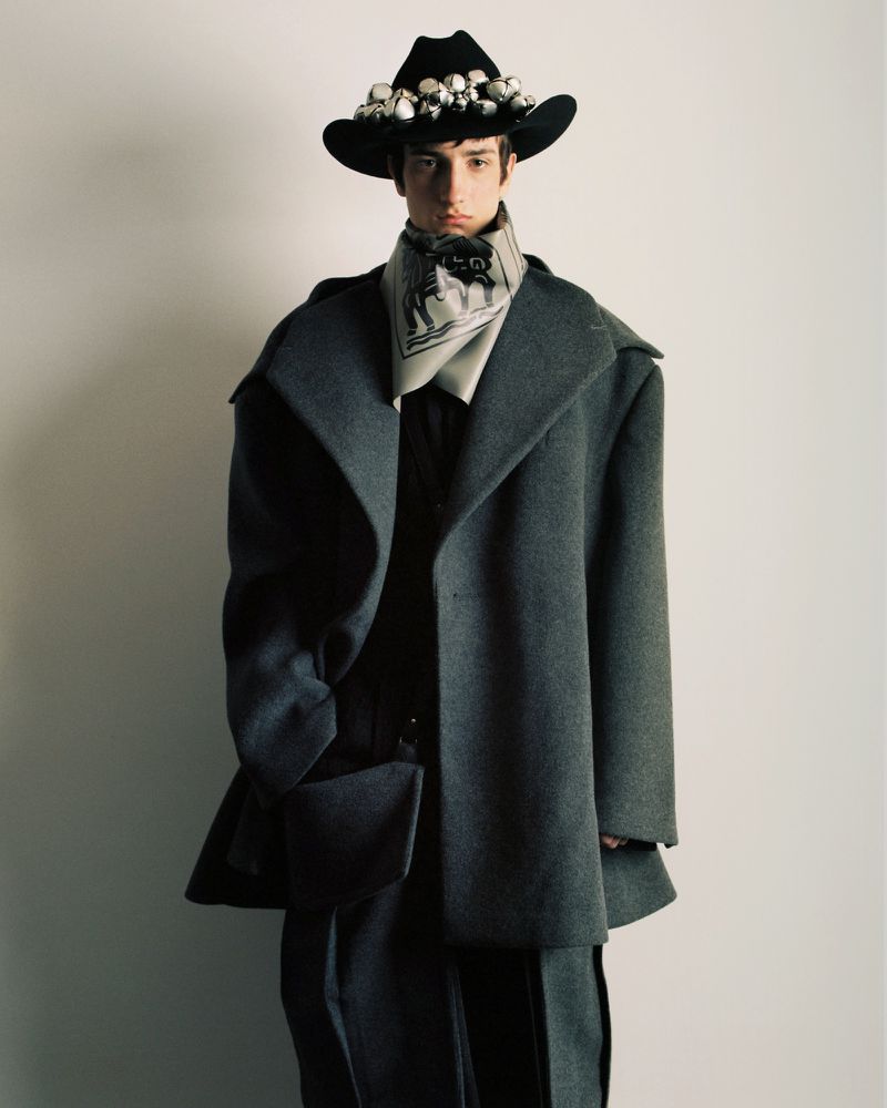Man in oversized grey coat and black cowboy hat adorned with white objects, posing against a white background in studio lighting.