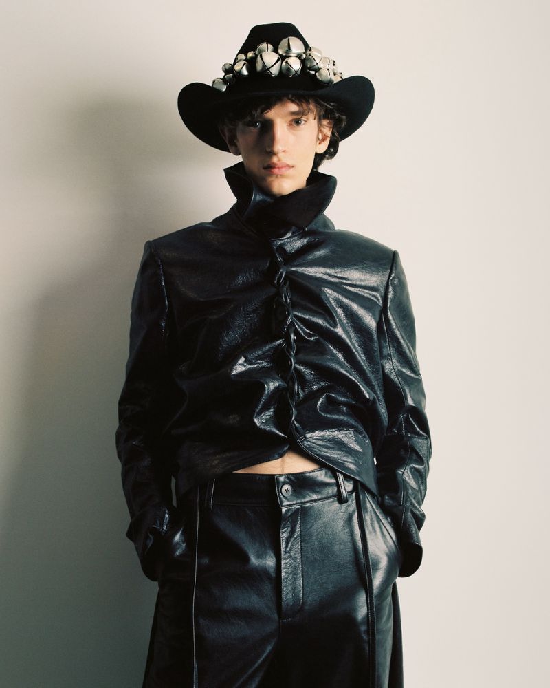 Male model in black shiny jacket and pants poses against a neutral wall, wearing a studded black cowboy hat for a fashion editorial.