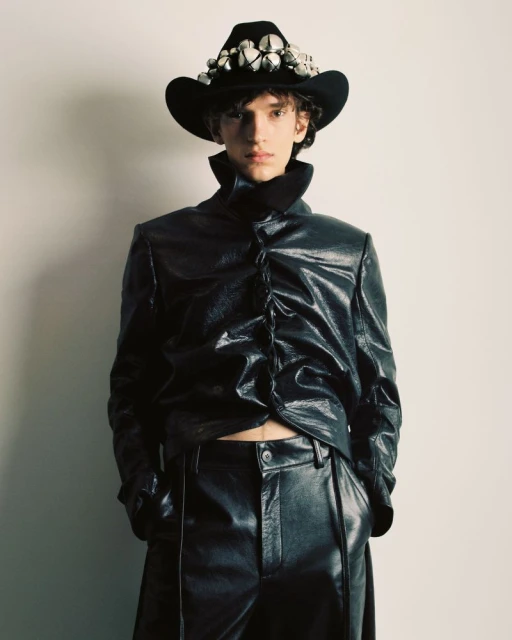 Male model in black shiny jacket and pants poses against a neutral wall, wearing a studded black cowboy hat for a fashion editorial.