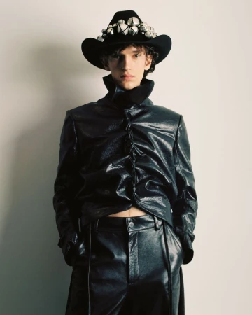 Male model in black shiny jacket and pants poses against a neutral wall, wearing a studded black cowboy hat for a fashion editorial.