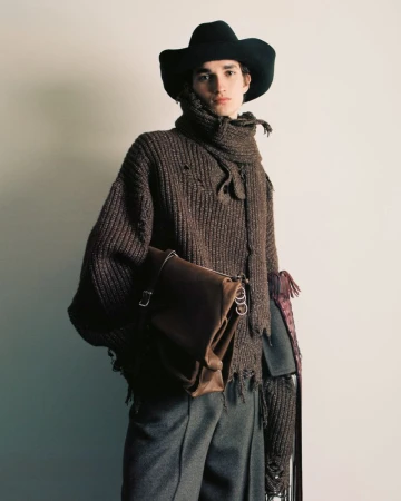 A model wearing a brown knit sweater, scarf, trousers and a hat stands indoors with a leather messenger bag in a fashion shot.