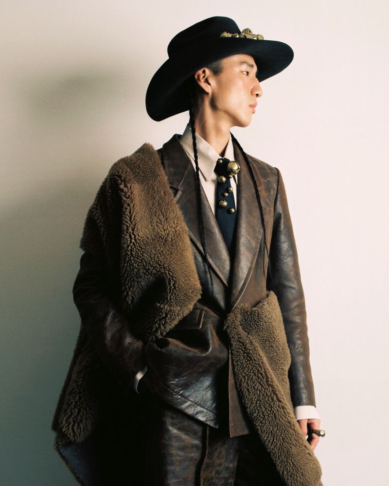 An Asian adult male wearing a black hat, brown suit, and tie poses in a photo studio with white background while glancing to the right.