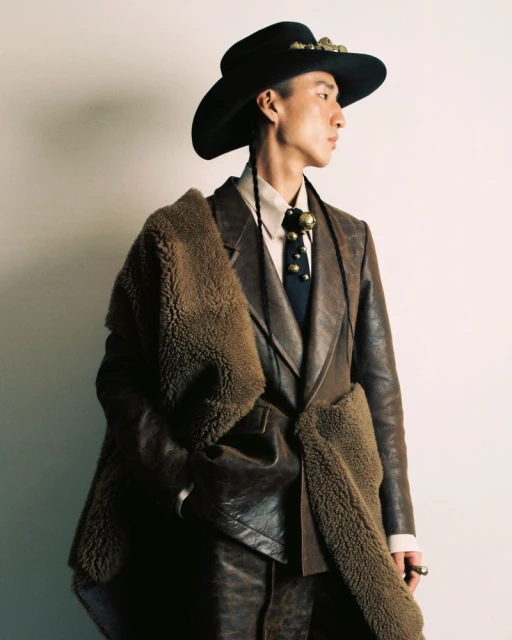An Asian adult male wearing a black hat, brown suit, and tie poses in a photo studio with white background while glancing to the right.