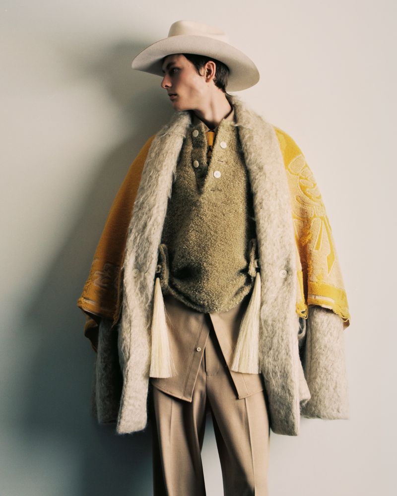 A model wears a cowboy hat, knit sweater, textured coat, and tailored pants in a studio shot against a neutral backdrop.