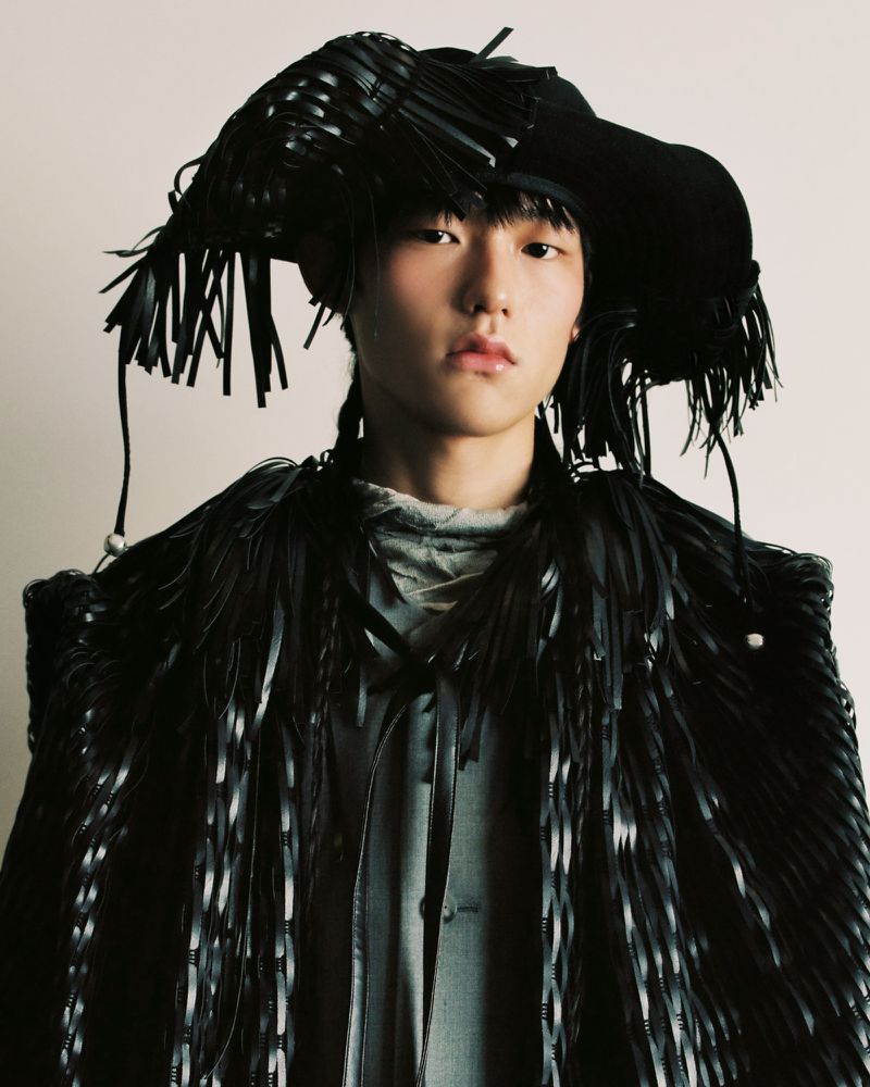 Portrait of a young adult posing in a black, fringed, avant-garde hat and coat, against a white backdrop in a fashion-forward style.