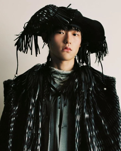 Portrait of a young adult posing in a black, fringed, avant-garde hat and coat, against a white backdrop in a fashion-forward style.
