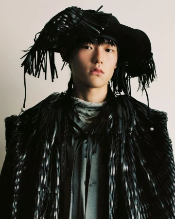 Portrait of a young adult posing in a black, fringed, avant-garde hat and coat, against a white backdrop in a fashion-forward style.