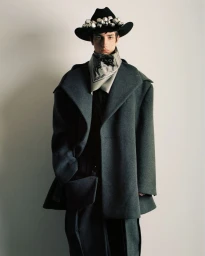 Model wearing a structured dark coat, printed scarf, and wide-brimmed hat with silver bells against a plain wall