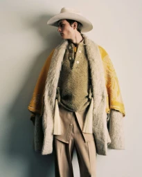 Model wearing a wide-brimmed hat, textured sweater, and layered coat against a plain background.