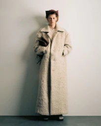 Model wearing a long textured cream coat and brown hat holding a dark bag against a plain grey studio backdrop.