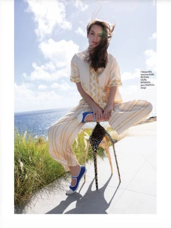 Model in a cream striped two-piece set and blue shoes sitting on a sculptural patterned stool overlooking the sea for Elle Bulgaria.