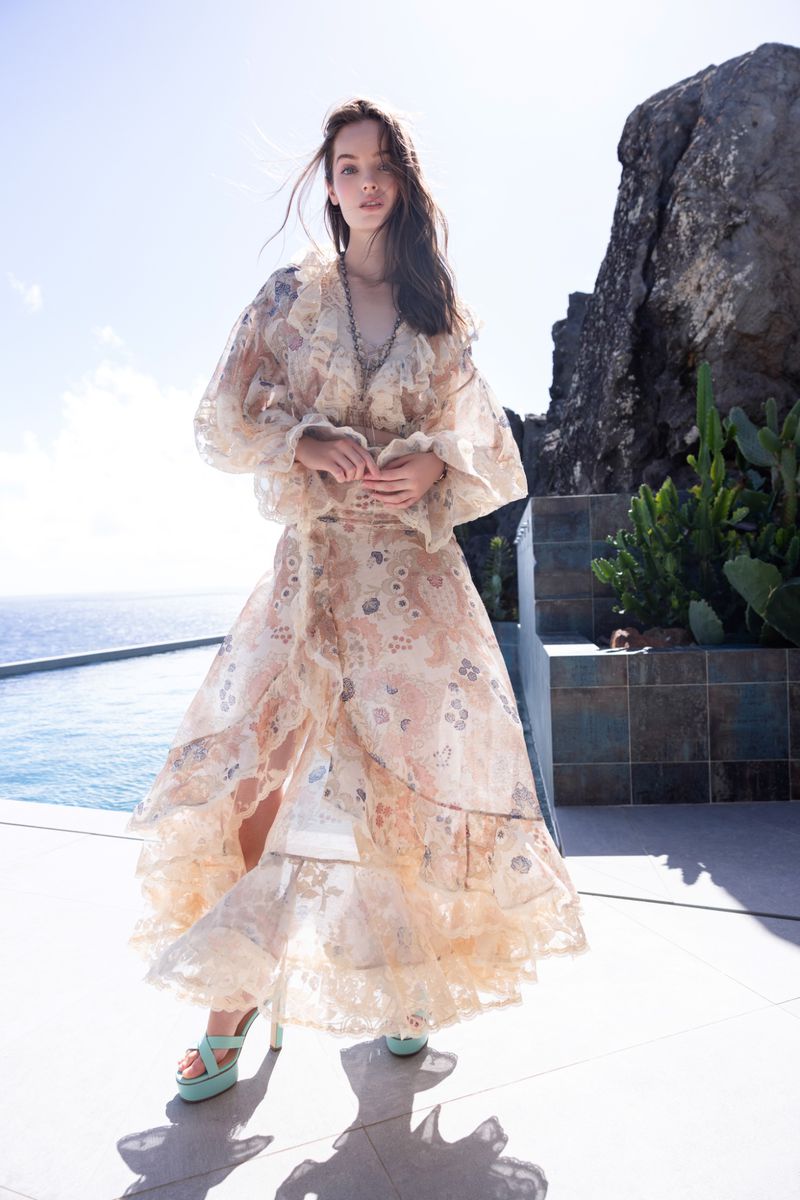 Model posing in a sheer ruffled floral gown and mint platform heels on an outdoor terrace by a pool overlooking the sea.