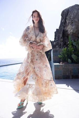Model posing in a sheer ruffled floral gown and mint platform heels on an outdoor terrace by a pool overlooking the sea.