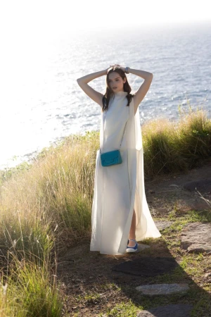 Model posing in a long sleeveless white dress and turquoise crossbody bag along a grassy coastal path overlooking the ocean.