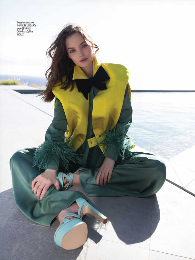 Fashion model seated outdoors by a pool in a green silk suit, bright yellow vest with black bow, and mint green platform heels.