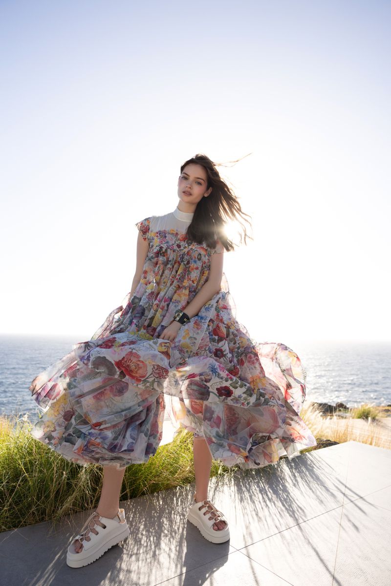 Model posing outdoors in a billowing sheer floral dress and white platform sandals against a bright sun and ocean horizon.