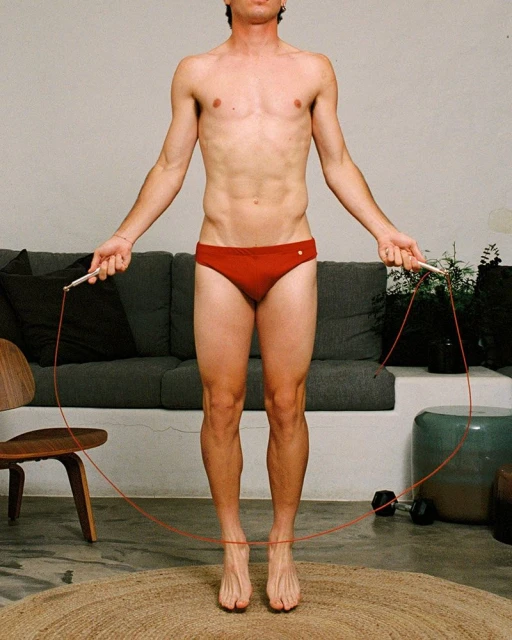 Athletic man in red swimsuit with jump rope indoors. Fitness and lifestyle shot in front of gray sofa and neutral background.