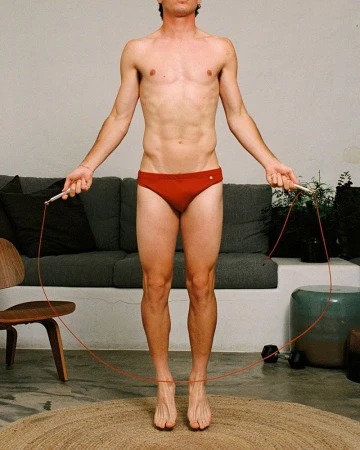 Athletic man in red swimsuit with jump rope indoors. Fitness and lifestyle shot in front of gray sofa and neutral background.