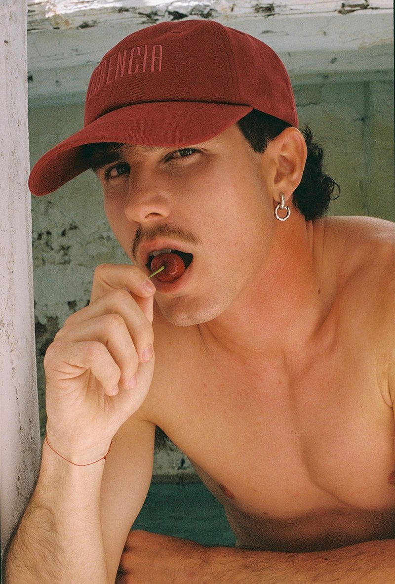 Attractive man in baseball cap poses indoors eating a cherry with bright lighting, hoop earring, and visible collarbone.