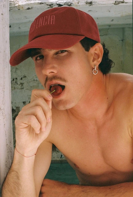 Attractive man in baseball cap poses indoors eating a cherry with bright lighting, hoop earring, and visible collarbone.