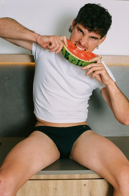 Young adult male in white shirt and black briefs vigorously bites into a large watermelon slice indoors with neutral background.