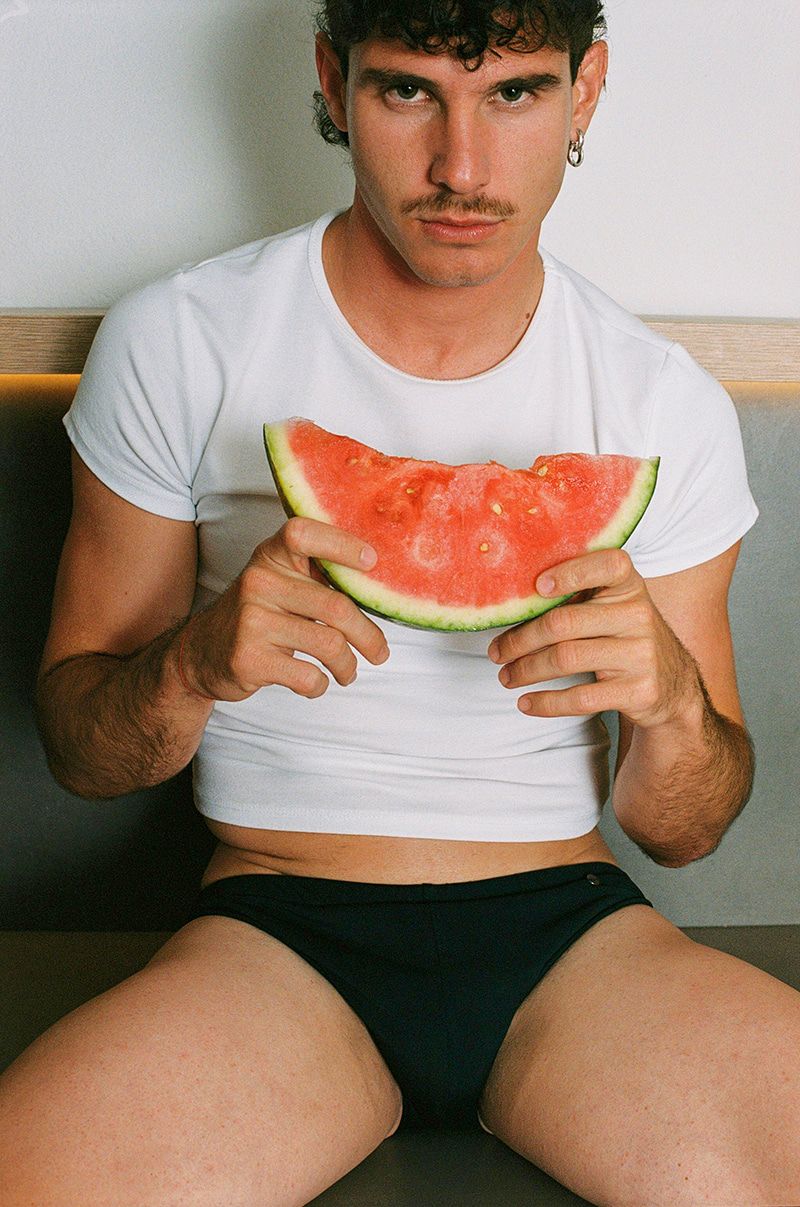 Young adult man in underwear holds a watermelon slice indoors against a neutral wall, showcasing summer fruit and healthy eating.