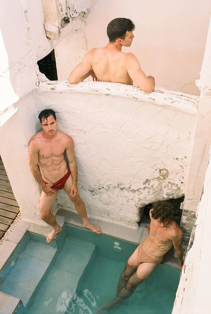 Three shirtless adult men in swimwear gather in a white, textured-walled pool. One is in the water while the others pose on the poolside.
