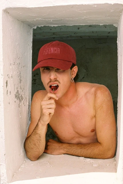 A young man with a cap in a window frame holds a cherry in his mouth. He is bare-chested in a rustic indoor setting with neutral lighting.