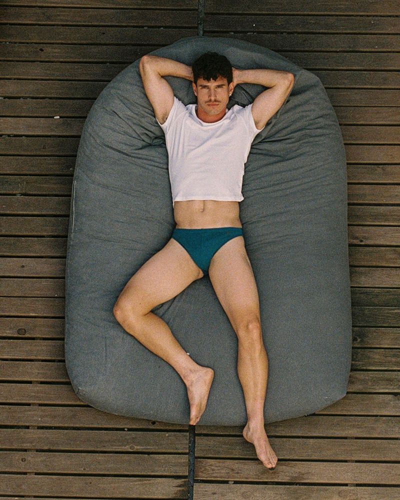 Athletic man lying on a gray beanbag cushion outdoors. He wears a white t-shirt and teal swimwear. Overhead shot of him on wood planks.
