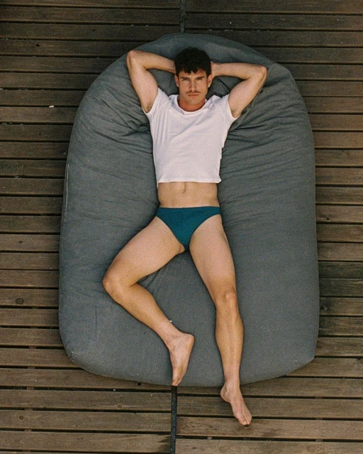 Athletic man lying on a gray beanbag cushion outdoors. He wears a white t-shirt and teal swimwear. Overhead shot of him on wood planks.