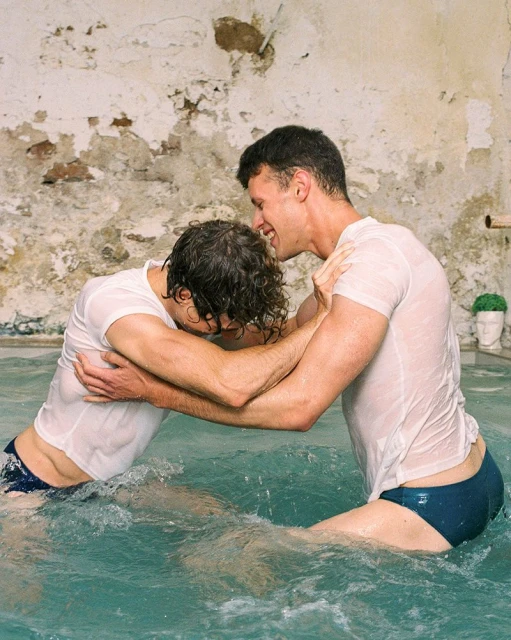 Two adult males are wrestling in turquoise water inside an old stone building. They are both wearing white t-shirts and dark swimwear.